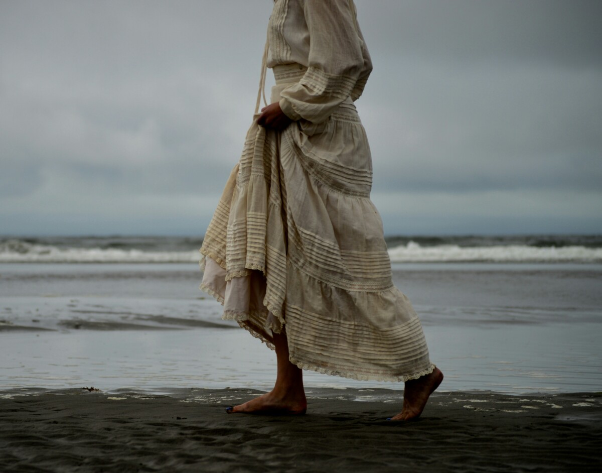 woman in white cotton dress walking along the shore of the ocean on a cloudy day